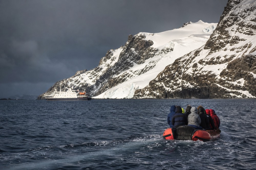 An orange inflatable boat carrying hooded parka-clad passengers pulls away from a white expedition ship anchored below a steep glaciated sub-Antarctic coastline