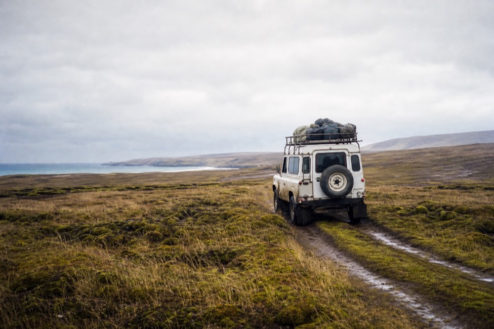 A mud-spattered white four-wheel drive crossing open moorland on a faint twin-track, low cloud over treeless hills, peat and tussock running to the horizon