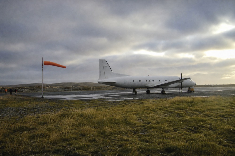 A white propeller aircraft parked on a windswept apron under flat grey cloud, an orange windsock stretched horizontal, empty moorland running to the fence line