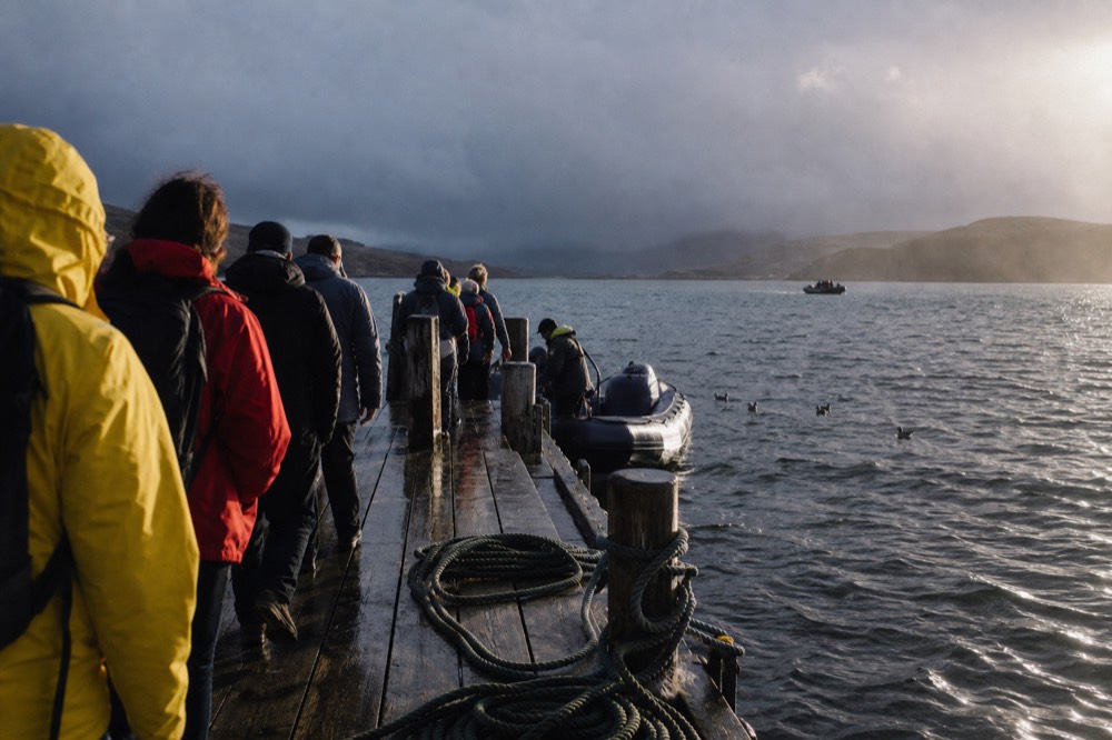 Passengers in waterproof jackets queuing along a wet wooden jetty in low afternoon light, coiled mooring ropes on the boards, gulls settled on the harbour water