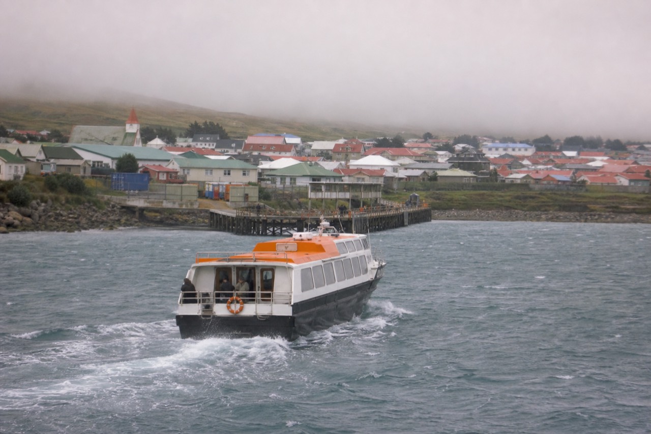 A cruise ship tender crossing grey chop toward the public jetty at Stanley, wind-flecked harbour water, painted metal roofs and low green hills behind the town