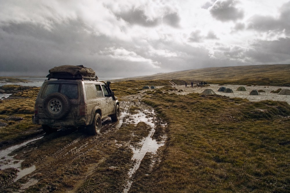 A mud-flanked 4x4 stopped on a rutted track over open camp, low sun catching standing water in the ruts, pale grass hills rolling away under heavy cloud