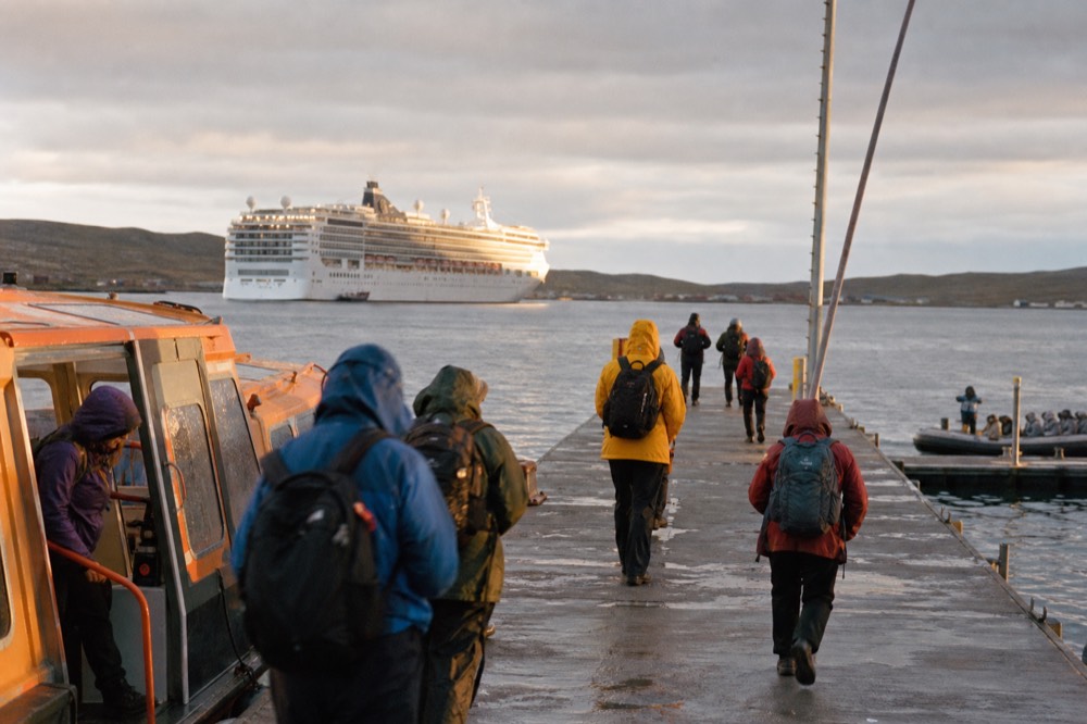 Passengers in waterproof jackets stepping from a small orange tender onto a concrete public jetty, a cruise ship at anchor in the harbour behind under grey sky