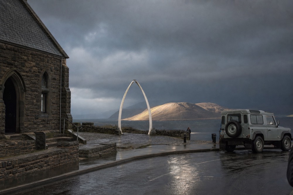 The whalebone arch outside a stone cathedral on a windswept waterfront street, wet tarmac reflecting pale light, a Land Rover parked at the kerb