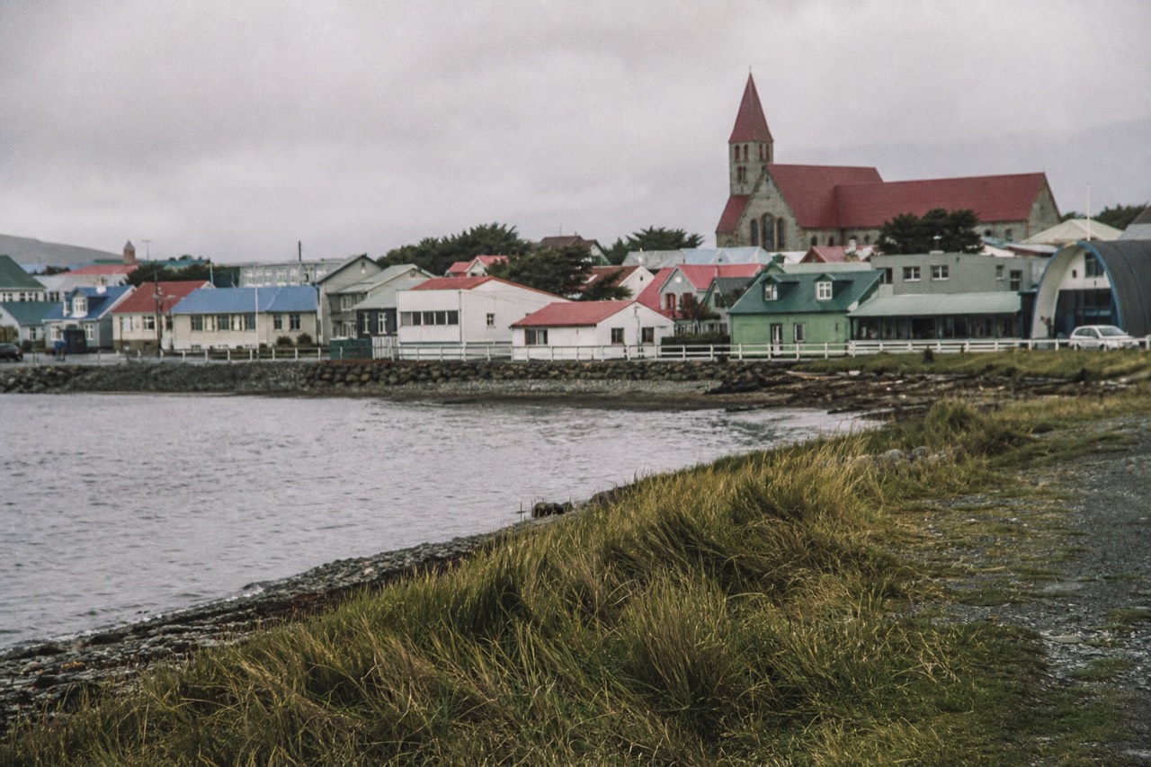 Stanley's waterfront row of small houses with brightly painted red green and blue metal roofs, the cathedral spire behind, grey harbour water and low cloud