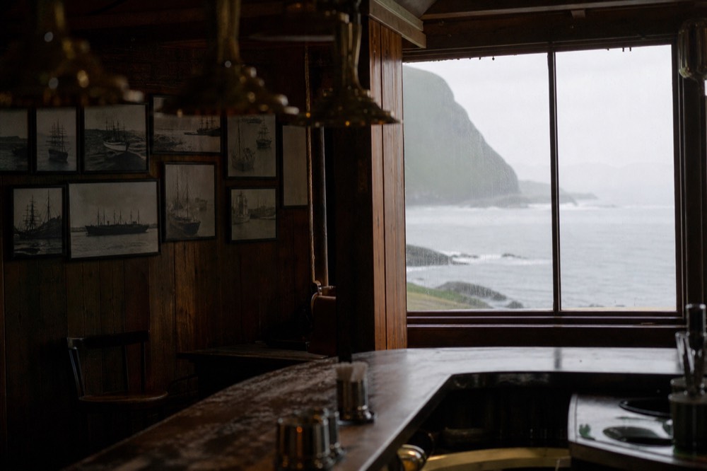 The dark wooden interior of a small island pub, brass fittings above a scuffed bar counter, framed ship photographs on the wall and rain streaking the window