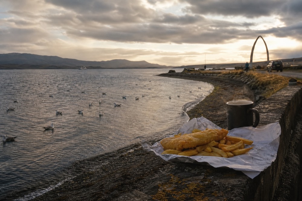 A paper wrap of golden fish and chips resting on a stone harbour wall beside a steaming mug, gulls on the grey water and a whalebone arch further along the shore