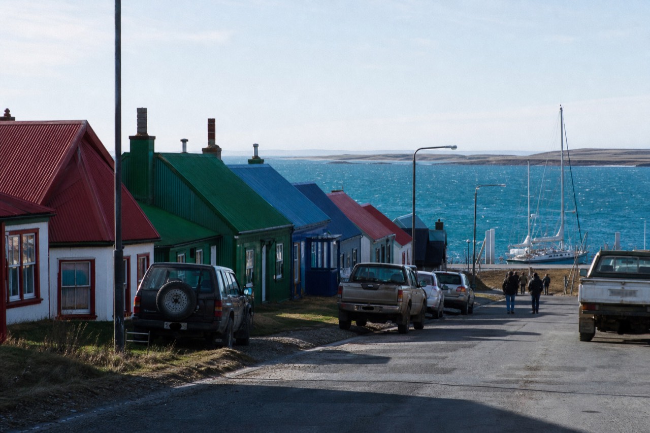 A row of brightly painted metal-roofed houses along the waterfront road in Stanley, red green and blue frontages, harbour water and a moored yacht behind them