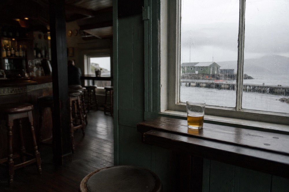 Interior of a small traditional pub, dark timber bar with brass fittings, mismatched wooden stools, rain-streaked window looking onto a grey harbour