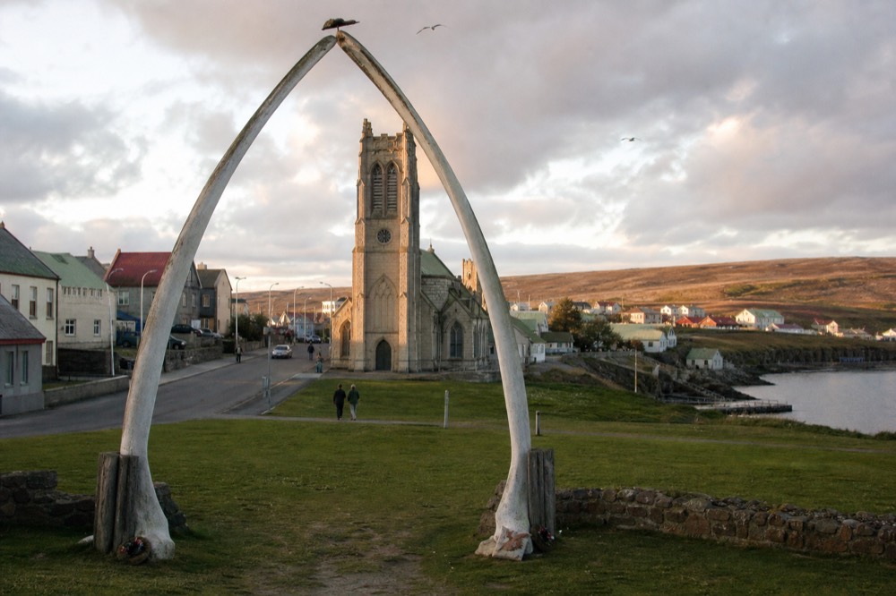 Whalebone arch framing a stone cathedral tower, cropped green lawn, low cloud, gulls overhead, an empty street running past behind