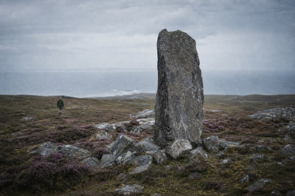 A single weathered stone memorial standing alone on open moorland, purple heath and scattered boulders around its base, distant sea and flat overcast light
