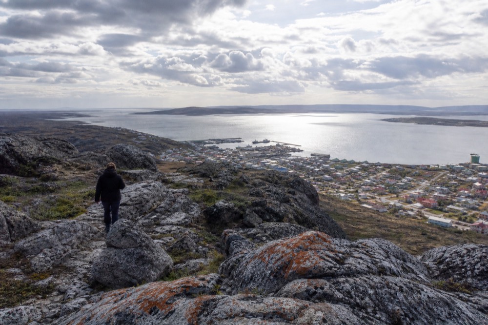 Rocky summit ridge above a small harbour town, lichen-streaked boulders in the foreground, coloured roofs and grey water visible far below in thin sunlight