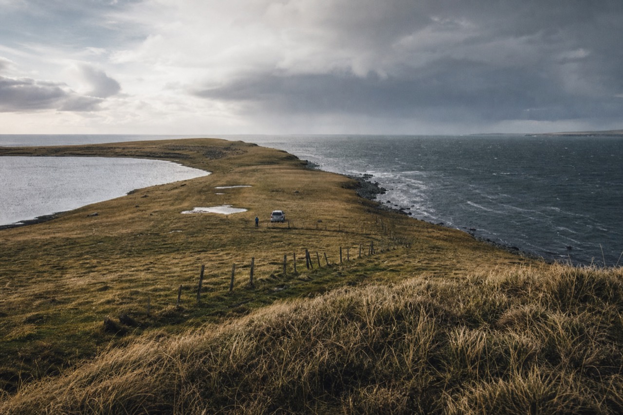 Narrow grass isthmus between two grey sea inlets seen from a low ridge, brown wind-flattened winter grass, sagging fence posts, heavy South Atlantic sky