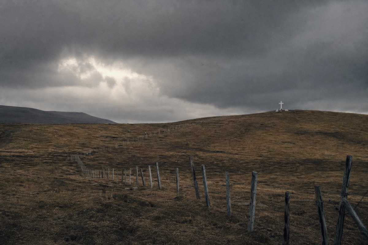 Rolling moorland at Goose Green with a fence line and memorial cross under a grey sky