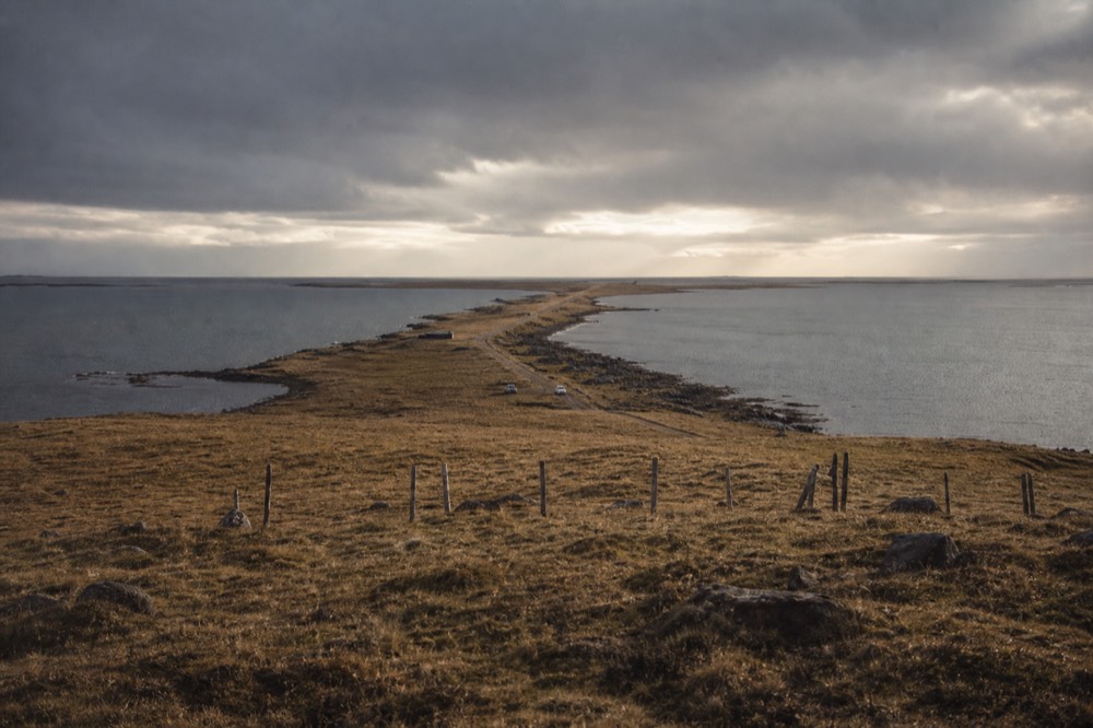 Narrow grass isthmus between two grey sea inlets seen from a low hill, wind-flattened winter grass, fence posts, heavy cloud pressing down