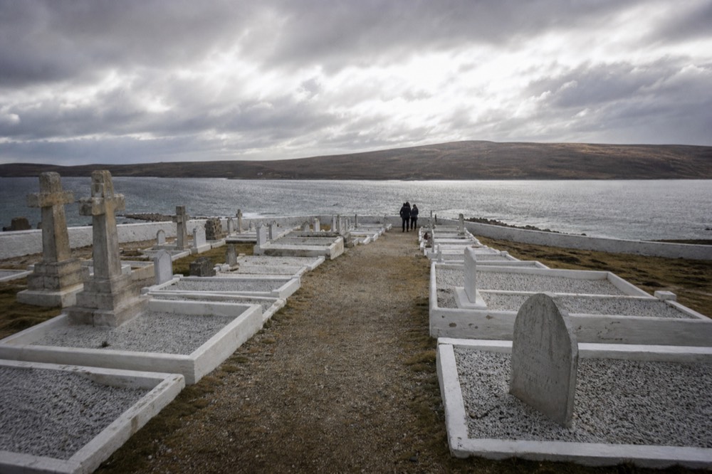 Rows of graves inside a low-walled cemetery enclosure on a windswept hillside, gravel path between them, sea inlet and bare moorland beyond under grey sky