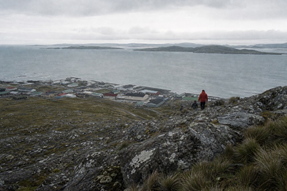 Rocky quartzite ridge above a small harbour town, lichen-covered boulders and scree in the foreground, painted roofs and grey water far below in thin light