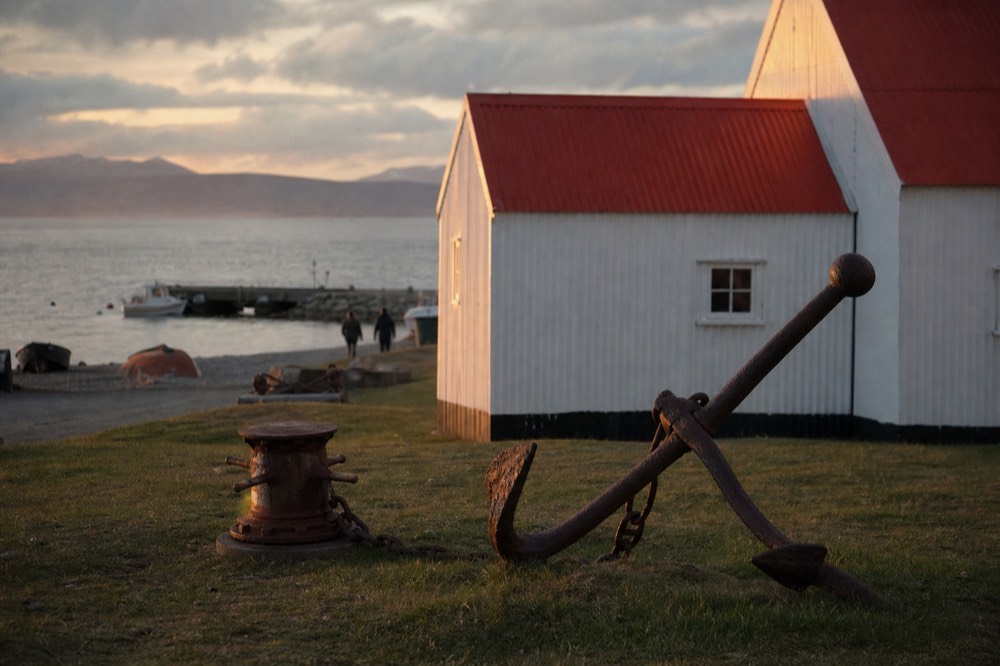 Whitewashed corrugated-iron museum buildings on a small waterfront, red roofs, a rusted anchor and old capstan set on cropped grass outside, grey harbour behind