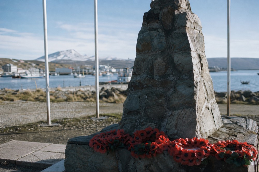 Stone memorial on a town waterfront with fresh poppy wreaths laid at its base, flagpole lines bare, grey harbour water and moored boats behind