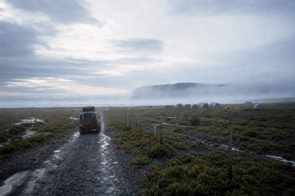 Four-wheel-drive vehicle on a rough gravel track crossing open camp, wire fence line running to the horizon, low scrub and standing water under fast-moving cloud