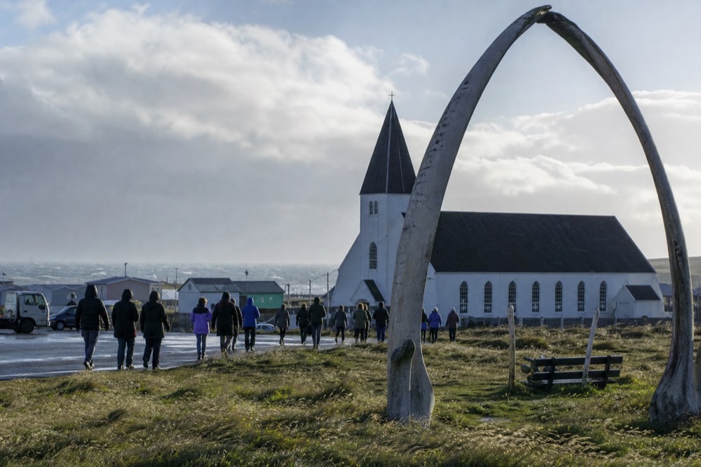 Busy small-town waterfront on a bright breezy morning with visitors in windproof jackets walking past a whalebone arch and a white weatherboard cathedral