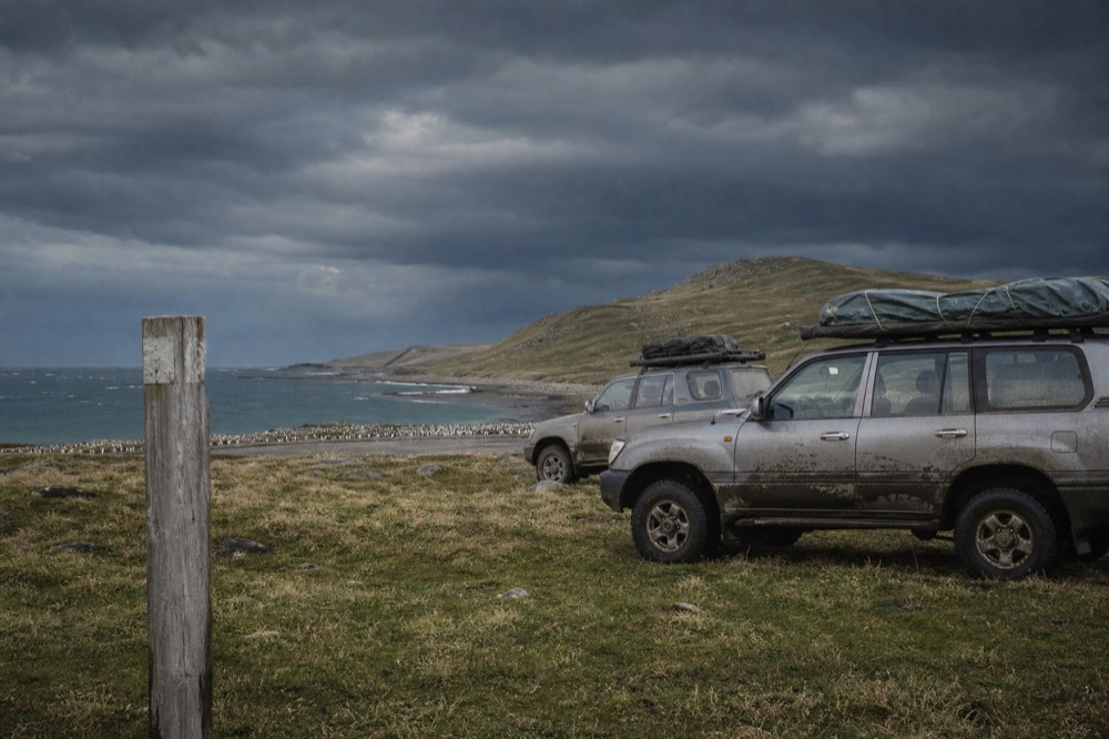 Convoy of two mud-streaked four-wheel drives parked on open camp beside a wooden marker post, king penguins visible as a pale band on the shore beyond