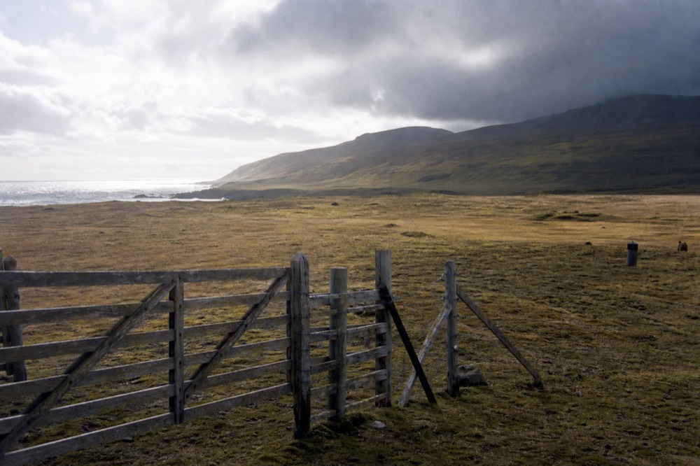 A wooden farm gate and stock fence standing on open camp grassland, fast cloud shadow moving across the hills behind