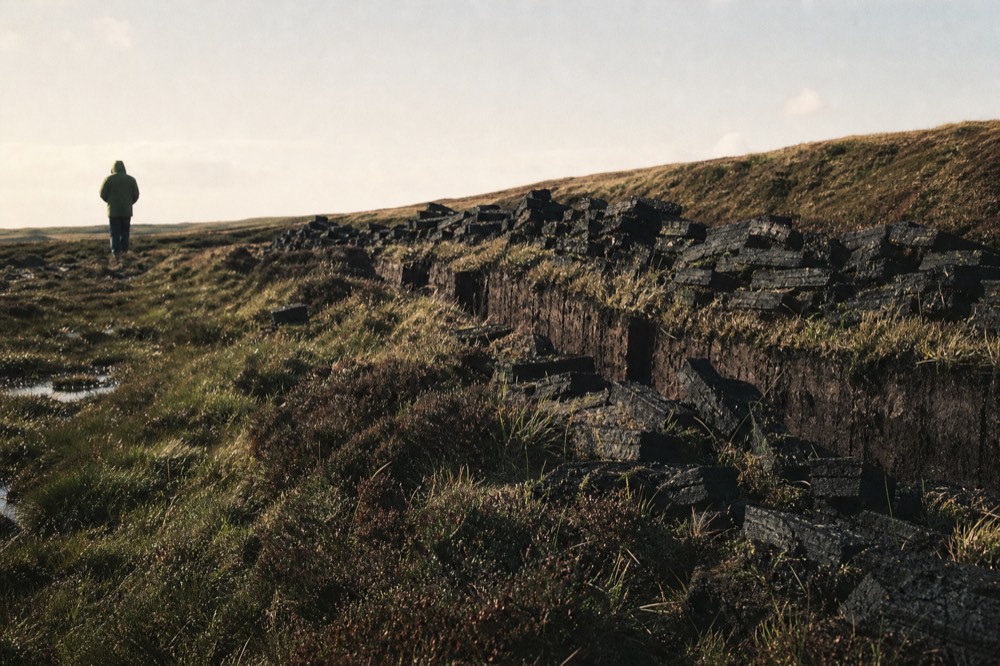 Peat banks cut into a moorland slope with dark stacked turves drying in the wind, heather and wet grass around them