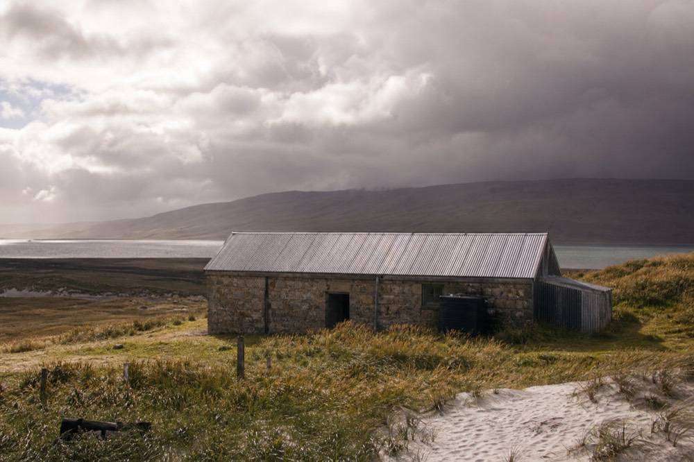 A small stone and corrugated-iron farm building alone on open grassland, dark weather closing in across the ridge behind it