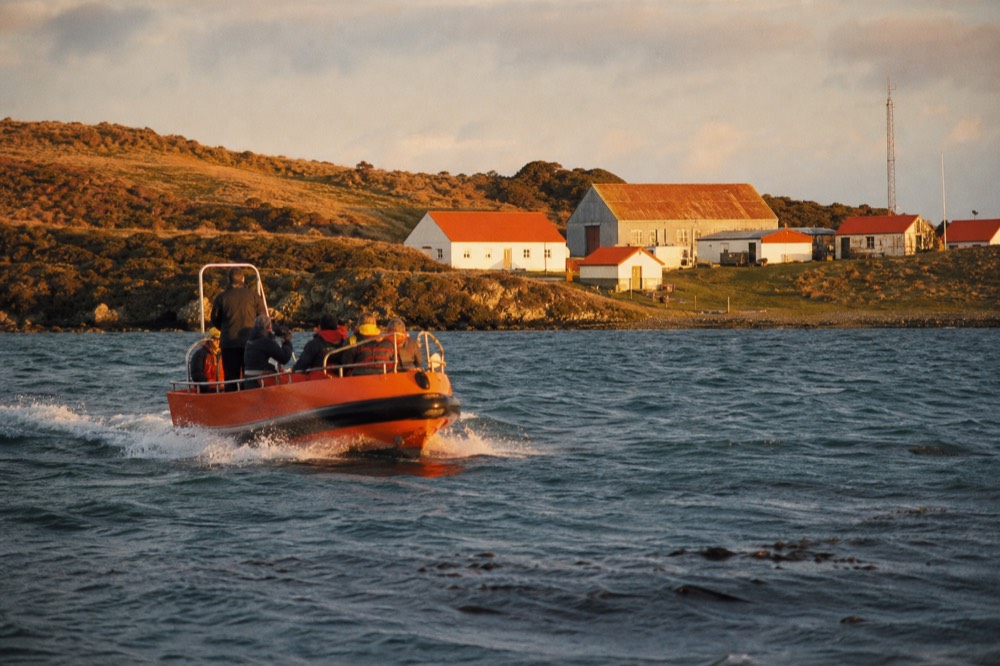Orange-hulled tender boat crossing choppy grey-green harbour water toward a low shoreline, white spray at the bow and dark kelp visible below the surface