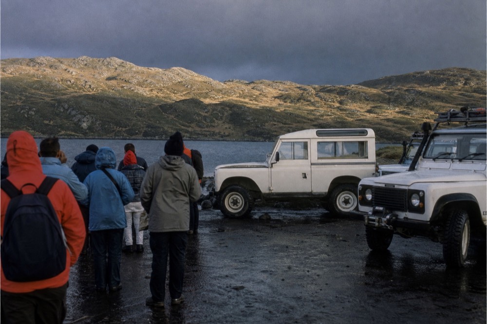 Small crowd of visitors in bright waterproof jackets gathered on a windy waterfront pavement beside parked white four-wheel-drive vehicles, harbour water and moorland behind
