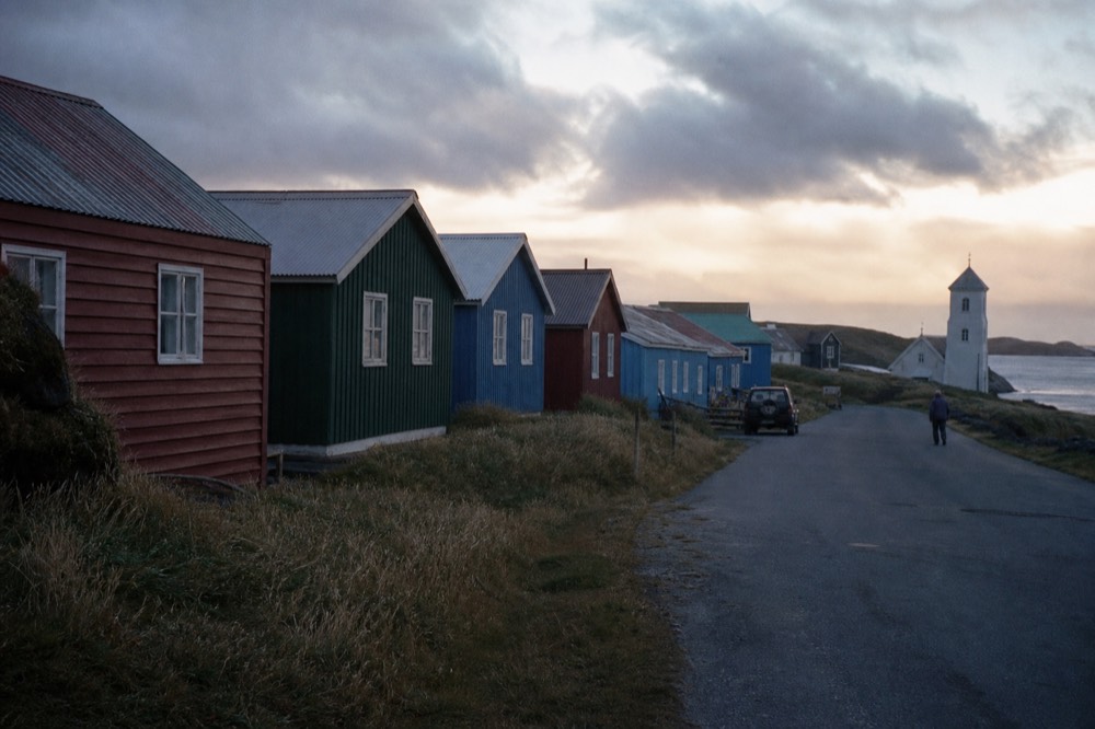 Row of small clapboard houses with corrugated roofs in deep red, green and blue along a quiet coastal road, a white weatherboard church tower rising at the far end