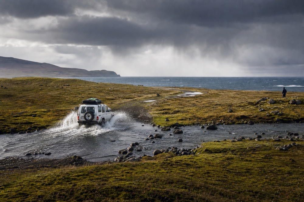 White four-wheel-drive vehicle throwing up spray as it crosses a shallow peat stream on an unmarked track over open camp, low hills and huge sky beyond