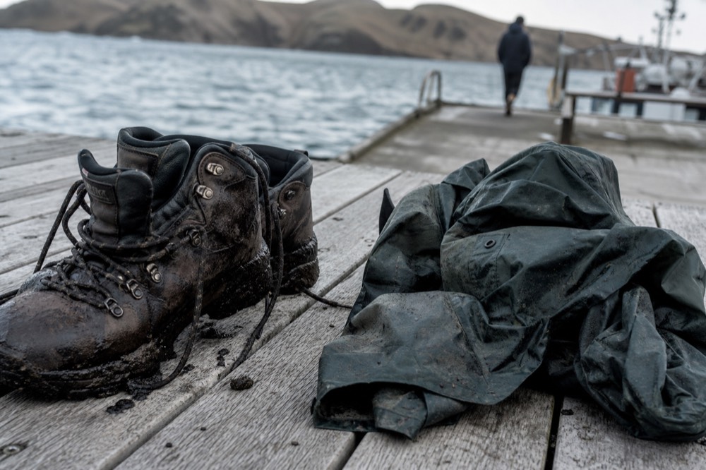 Scuffed leather walking boots caked in dark peat mud beside a folded waterproof shell on weathered decking, drying in flat southern daylight