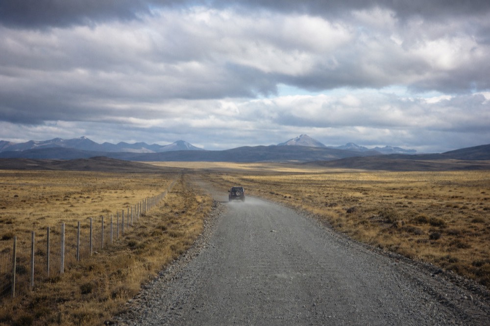 Long straight gravel road cutting across empty pale-gold moorland toward distant blue-grey ridges, a single fence line running parallel under scudding cloud
