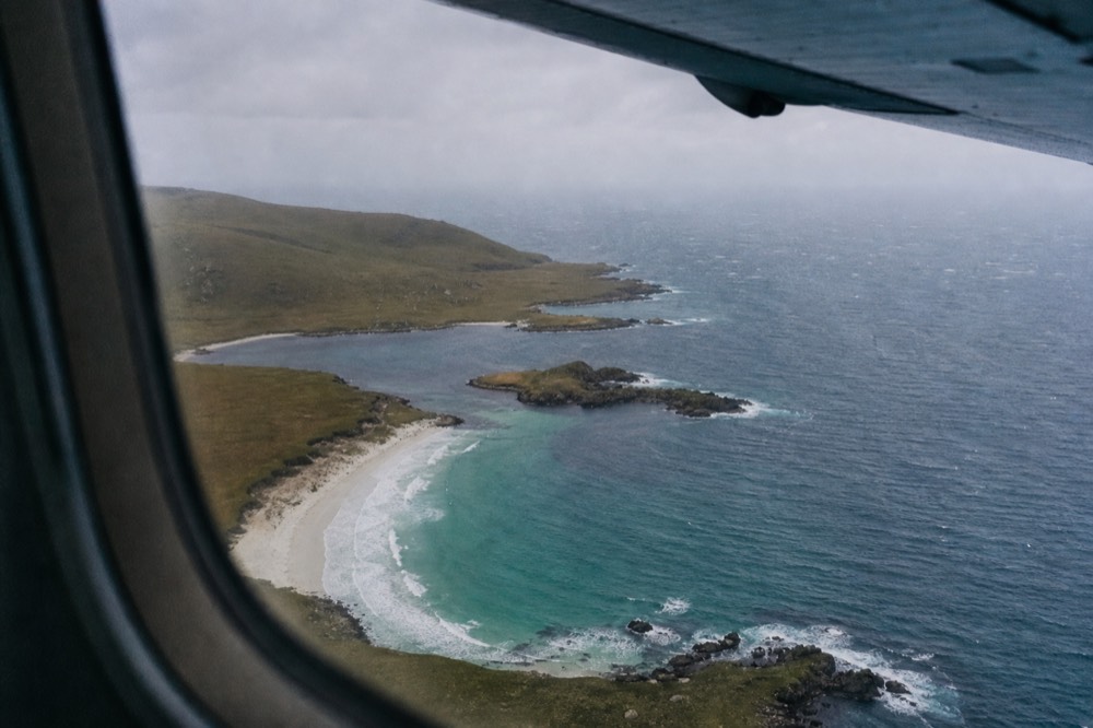 View through a small aircraft window over a broken coastline of dark headlands and turquoise shallows, white surf lines curving around an empty sandy bay
