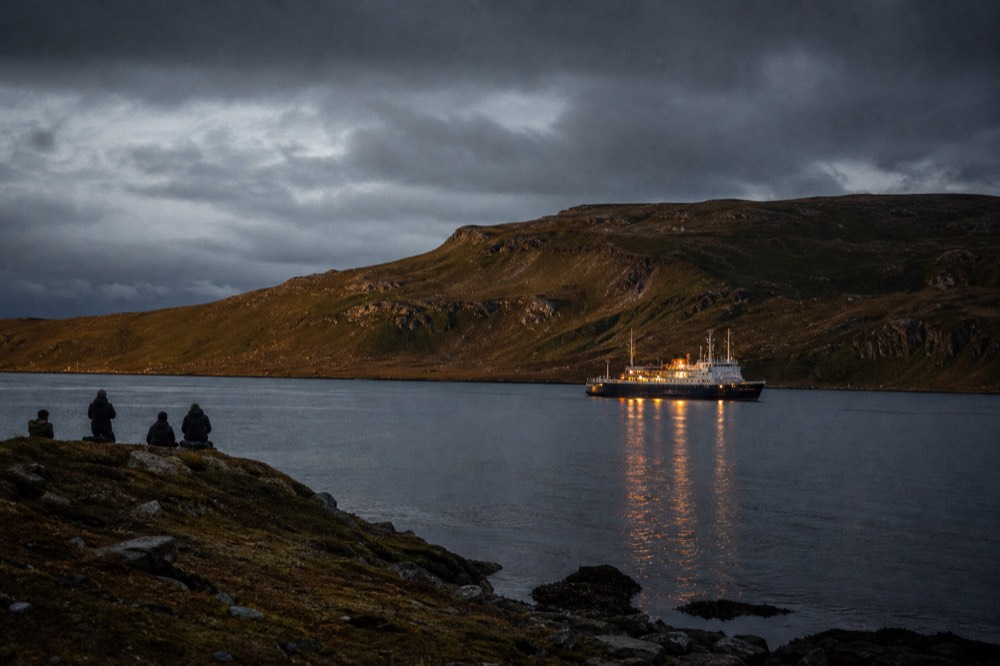 Expedition ship lying at anchor in a wide sheltered bay at dusk, deck lights reflected on flat pewter water with dark low hills wrapping the far shore