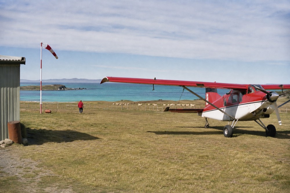 Small red and white Islander aircraft on a grass airstrip with a windsock and a single shed, sheep grazing beyond a wire fence under high thin cloud