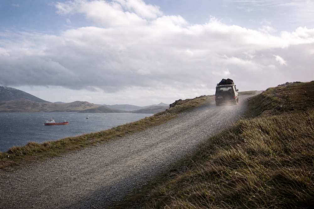 A gravel road climbing a bare headland above a harbour, wind-flattened grass on both verges, sea visible beyond the crest