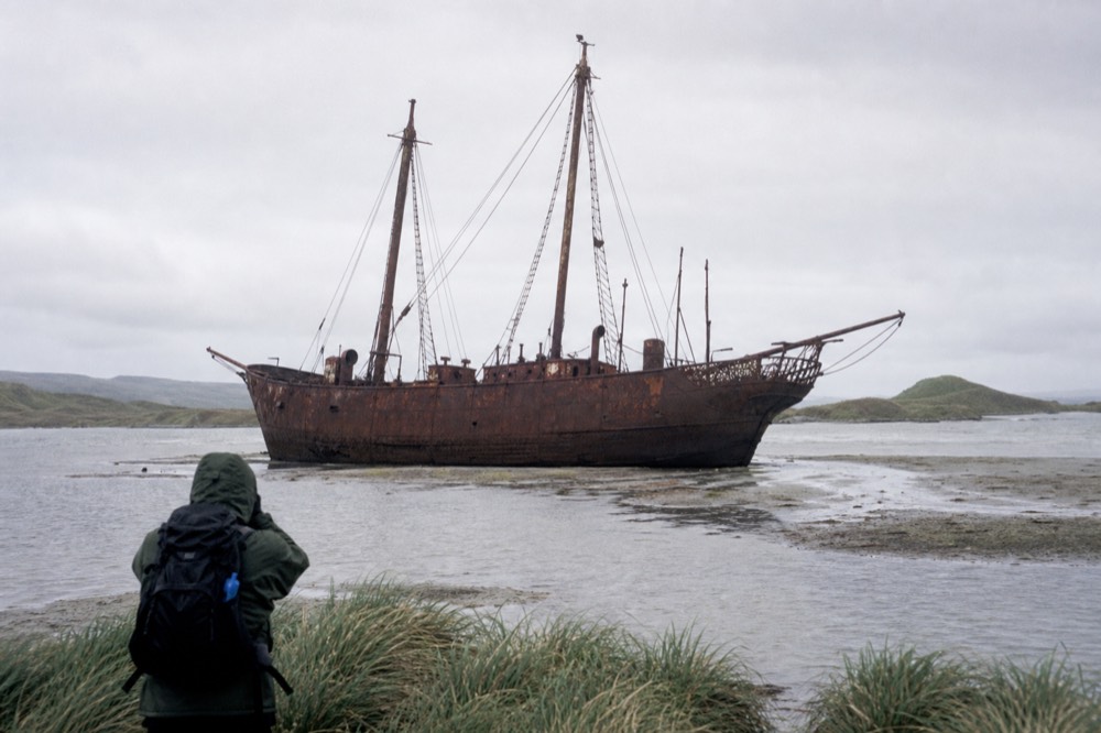 A rusted iron sailing-ship hulk sitting upright on a sandbank in shallow water at low tide under a flat grey sky
