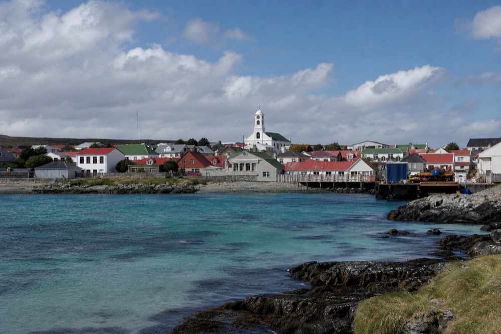 Stanley's painted metal roofs in ranked rows above a waterfront, a white cathedral tower rising among them under bright broken cloud