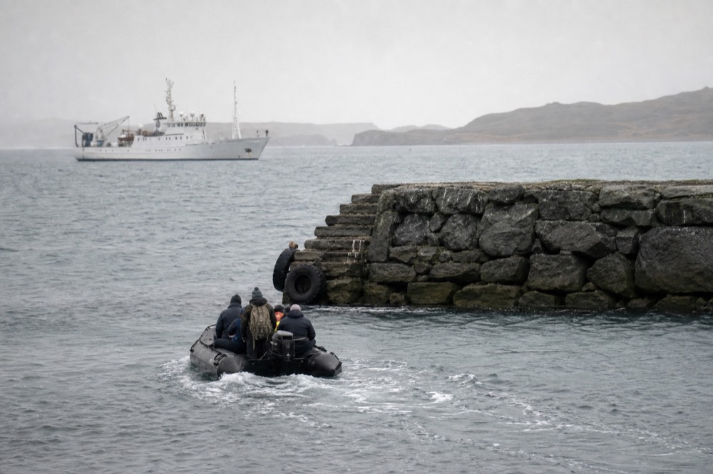 A small tender boat approaching a stone jetty in a sheltered harbour, a white ship anchored offshore on flat grey water