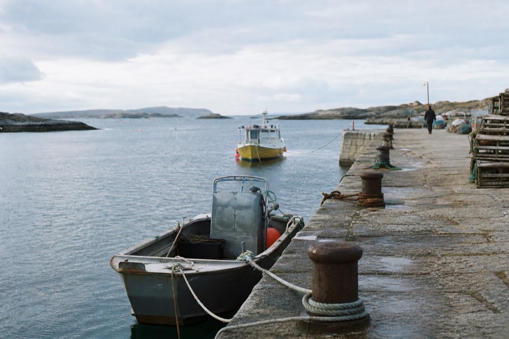 Stone jetty with iron bollards and coiled mooring rope, small workboats tied alongside in flat grey water on a still morning