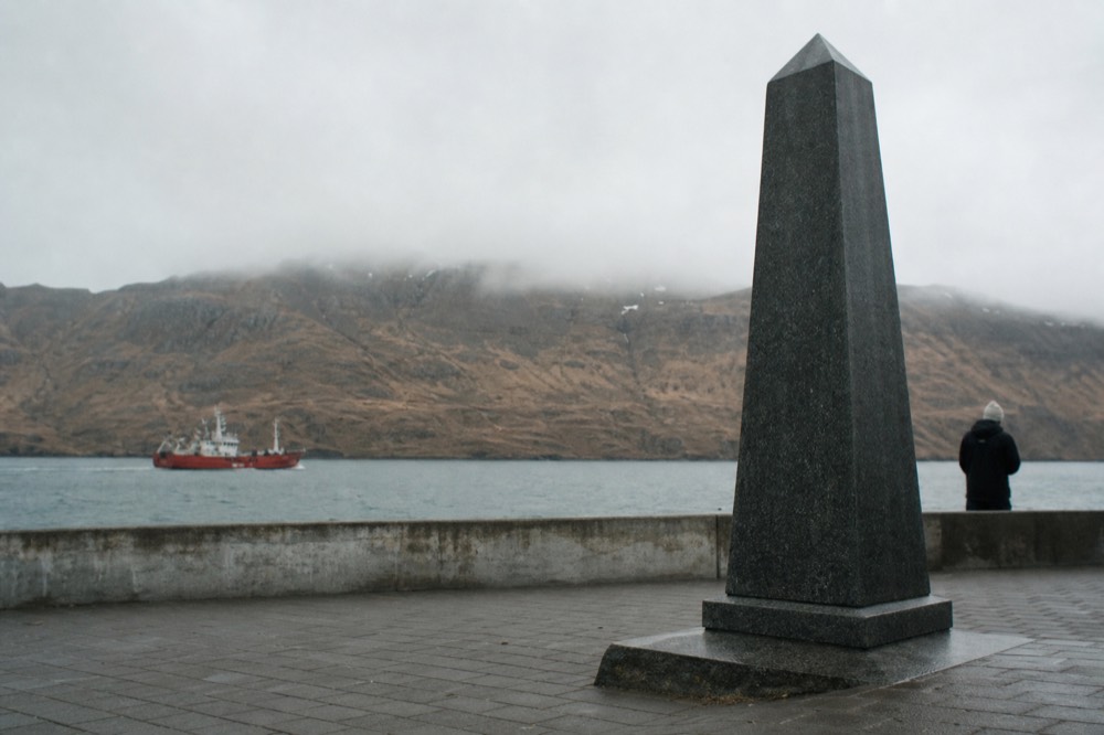Tall dark stone memorial standing on a paved harbour front terrace with a low wall, grey sea and bare brown hills behind it