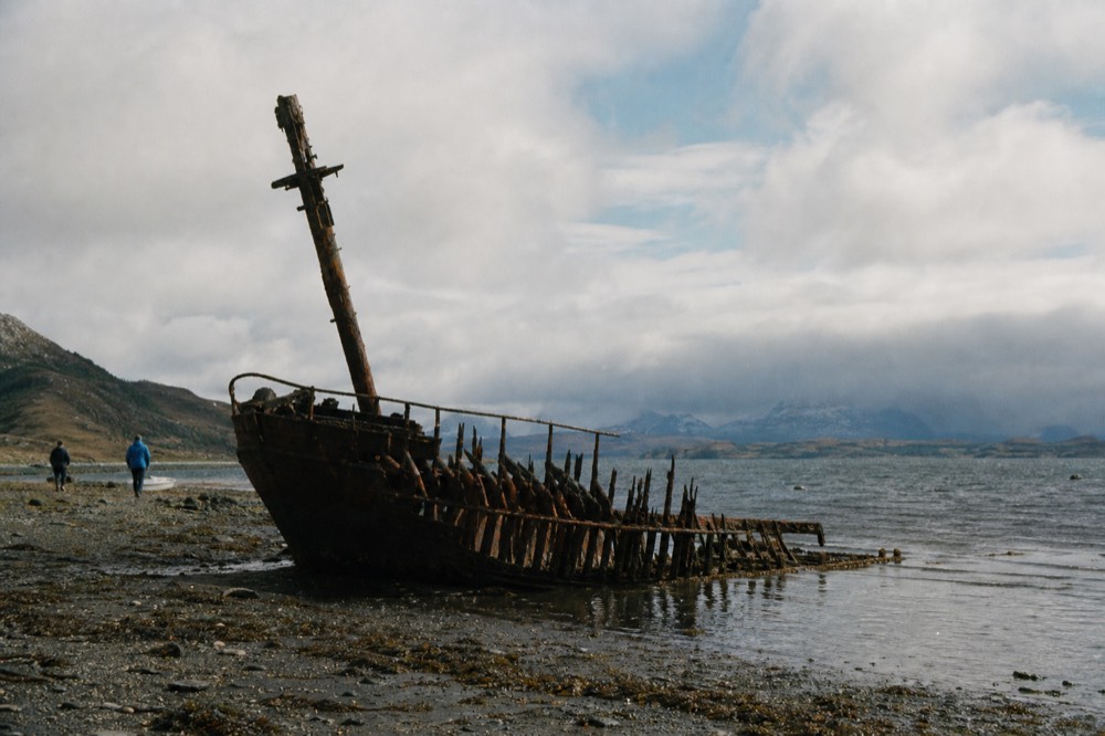 Rusted iron hull of an old sailing ship half sunk in shallow harbour water at low tide, exposed ribs dark against a pale sky