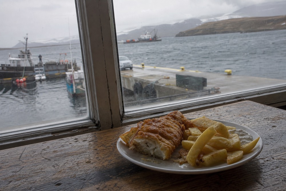 Plate of battered fish and thick cut chips on a scrubbed wooden table beside a window looking out onto a grey harbour