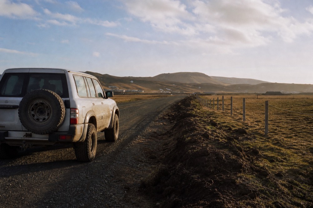 Mud splashed white four wheel drive parked on a gravel road at the edge of town, peat banks and a wire fence running toward low brown hills