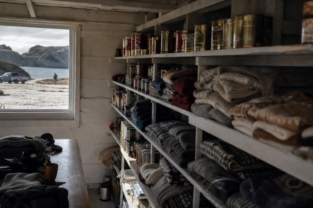 Shelves of tinned supplies and folded hand knitted wool goods inside a small island general store lit by a bare window