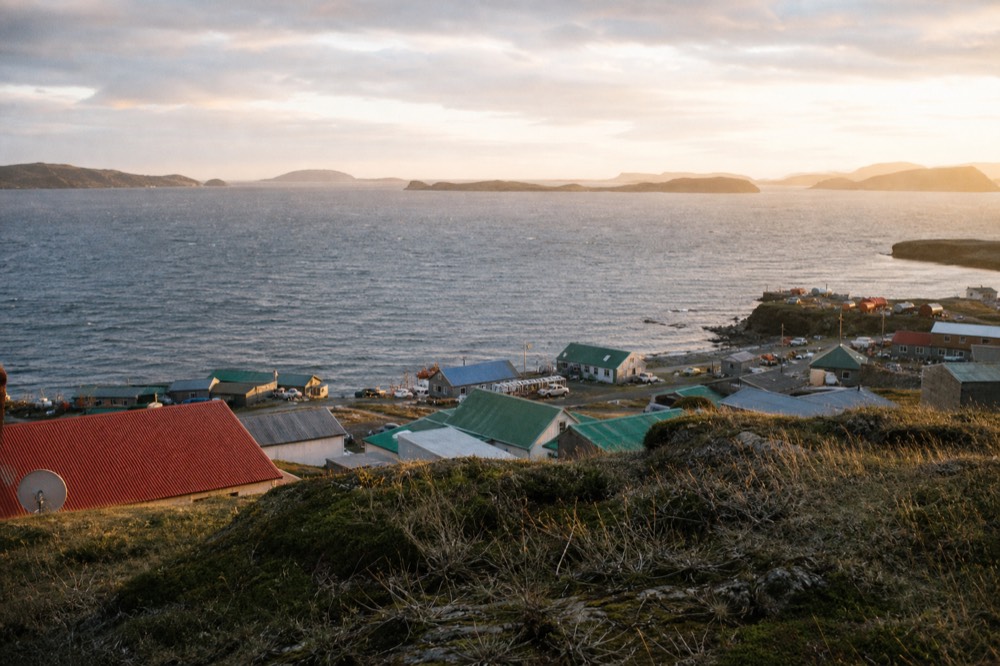 Painted metal roofs of a small harbour town in green, red and blue, seen from a hillside above a wind-ruffled inlet
