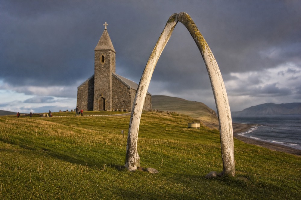 Weathered arch made from two pale whale jawbones standing on cropped grass in front of a stone church tower under bright southern light